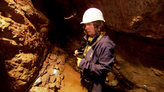 Die Bergwerksfrau steht mit einer Taschenlampe in einem dunklen Stollen der Grube Sophia und leuchtet die feuchten Felswände des historischen Bergwerks aus.