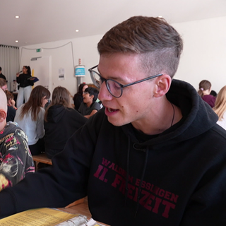 Noah sitzt an einem Tisch und spielt ein Spiel, während im Hintergrund viele Kinder mitspielen und das lebendige Ferienlager sichtbar wird.