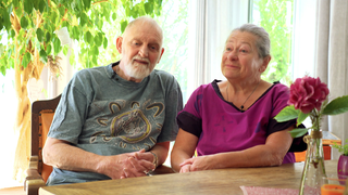 Michael und Ulrike sitzen nebeneinander an einem Tisch, Michael erzählt lebhaft eine Geschichte, während Ulrike ihm aufmerksam zuhört.