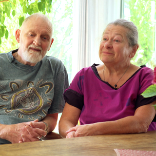 Michael und Ulrike sitzen nebeneinander an einem Tisch, Michael erzählt lebhaft eine Geschichte, während Ulrike ihm aufmerksam zuhört.