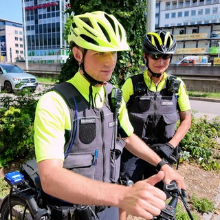 Ein Fahrradpolizist und eine Fahrradpolizistin sind in Stuttgart unterwegs. Sie stehen an einer Straße.