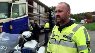 Verkehrskontrolle der Autobahnpolizei auf der A81: ein LKW-Fahrer wird kontrolliert. Im Vordergrund steht ein Polizist in gelber Warnweste.