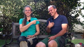 Raphaela und Cornelius Grauer sind seit 20 Jahren ein Paar. Sie sitzen auf einer Holzbank im Garten und beide zeigen mit dem Finger auf Cornelius.