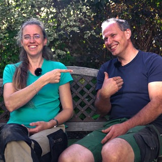 Raphaela und Cornelius Grauer sind seit 20 Jahren ein Paar. Sie sitzen auf einer Holzbank im Garten und beide zeigen mit dem Finger auf Cornelius.