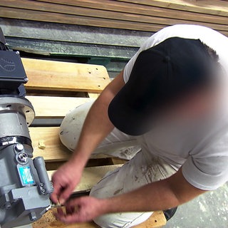 Insasse der JVA Mannheim arbeitet im Malerbetrieb. Er kniet auf einer Palette und arbeitet an einer Maschine.