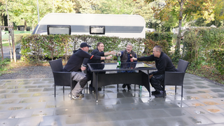 Vier Männer stoßen auf eine schöne Campingsainson an. Sie sitzen an einem Tisch und prosten sich zu. Im Hintergrund ein Camper. 