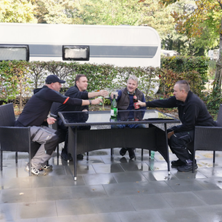 Vier Männer stoßen auf eine schöne Campingsainson an. Sie sitzen an einem Tisch und prosten sich zu. Im Hintergrund ein Camper. 