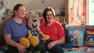 Sonja Schreiber und Ottmar Gamma sitzen zusammen auf einem Sofa. Zwischen ihnen eine große Euromaus, das Maskottchen des Europaparks. 