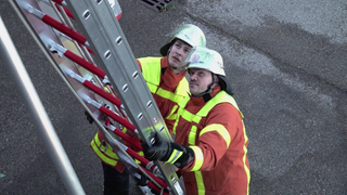 Zwei Feuerwehrmänner halten eine Leiter und schauen nach oben. Sie sind Teil einer Feuerwehrübung.