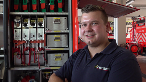 Ein Mann steht vor einem Feuerwehrauto. Er trägt ein Polo-Shirt mit der Aufschrift: Feuerwehr. Das Fahrzeug ist geöffnet.