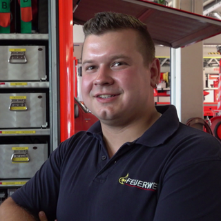 Ein Mann steht vor einem Feuerwehrauto. Er trägt ein Polo-Shirt mit der Aufschrift: Feuerwehr. Das Fahrzeug ist geöffnet.