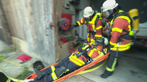 Drei Feuerwehrmänner in Einsatzkleidung tragen einen weiteren Feuerwehrmann auf einer Trage.