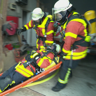 Drei Feuerwehrmänner in Einsatzkleidung tragen einen weiteren Feuerwehrmann auf einer Trage.