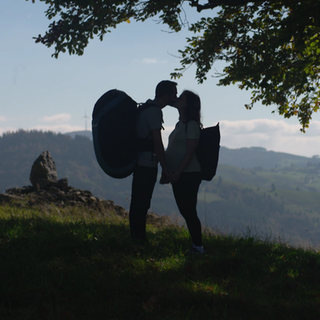 Ein Paar küsst sich. Er trägt einen großen Rucksack, in dem ein Gleitschirm ist. Sie ist schwanger.