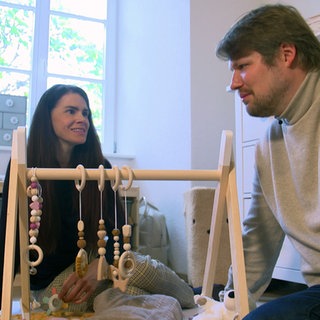Bürgermeisterin Jessica Lang und Bürgermeister Philipp sitzen in einem Zimmer neben ihrem Baby, das auf einer Decke liegt.