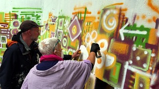 Graffiti-Workshop in Mannheim: Ein Mann mit Kappe und eine Frau mit grauem Haar besprühen Wand.