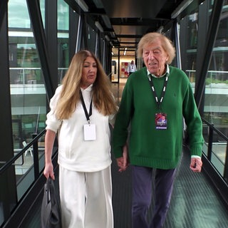 Olga und Bata Illic halten sich an den Händen und laufen durch einen gläsernen Flur.