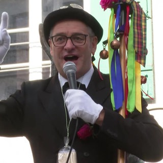 Ein "Fasnetslader" spricht mit Anzug, Hut und Mikro zu den Besuchern