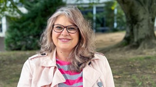 Susanne Sarantis trägt eine rundliche Brille, trägt einen Nasenpiercing und lächelt freundlich mit Zähnen in die Kamera.