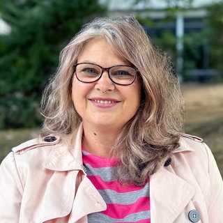 Susanne Sarantis trägt eine rundliche Brille, trägt einen Nasenpiercing und lächelt freundlich mit Zähnen in die Kamera.