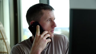 Ultraläufer Daniel Hernes sitzt an einem Computer im Büro. Er hat ein Handy am Ohr und telefoniert.