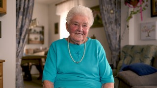 Rösle sitzt in ihrem Wohnzimmer. Sie trägt ein türkisfarbenes T-Shirt und eine Perlenkette. Sie hat kurze, graue Haare und lacht in die Kamera.