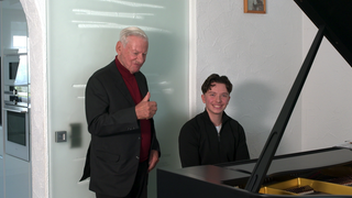 Ein junger Mann sitzt an einem Konzertflügel. Neben ihm steht ein älterer Herr und streckt seinen Daumen nach oben. Beide lächeln. 