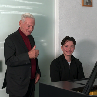 Ein junger Mann sitzt an einem Konzertflügel. Neben ihm steht ein älterer Herr und streckt seinen Daumen nach oben. Beide lächeln. 