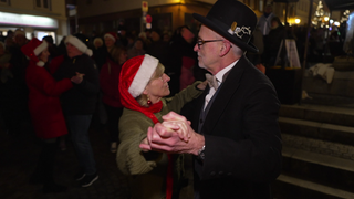 Ein Paar tanz auf dem Weihnachtsmarkt. Sie trägt eine lange Weihnachtsmannmütze, er einen Anzug und einen schwarzen Hut. Im Hintergrund sind viele tanzende Paare.
