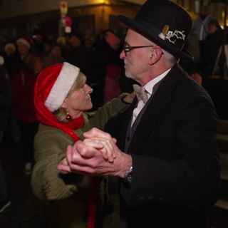 Ein Paar tanz auf dem Weihnachtsmarkt. Sie trägt eine lange Weihnachtsmannmütze, er einen Anzug und einen schwarzen Hut. Im Hintergrund sind viele tanzende Paare.