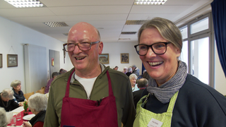 Hansi Gerber und seine Frau Anja lächeln. Er trägt eine rote Schürze, sie eine grüne. Im Hintergrund sitzen Senioren an einem Tisch. 