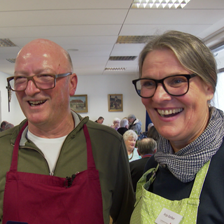 Hansi Gerber und seine Frau Anja lächeln. Er trägt eine rote Schürze, sie eine grüne. Im Hintergrund sitzen Senioren an einem Tisch. 