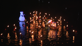 In einem Fluss schwimmen Menschen in Neoprenanzügen. Jeder hält eine Fackel über der Wasseroberfläche. Ein mit Luft gefüllter, schwimmender Schneemann treibt auf dem Fluss.  