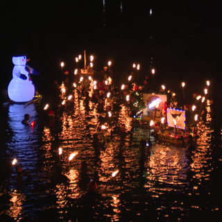 In einem Fluss schwimmen Menschen in Neoprenanzügen. Jeder hält eine Fackel über der Wasseroberfläche. Ein mit Luft gefüllter, schwimmender Schneemann treibt auf dem Fluss.  