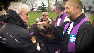 Ein Pfarrer streichelt einen Hund am Kopf, der bei seinem Besitzer auf dem Arm ist.