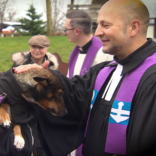 Ein Pfarrer streichelt einen Hund am Kopf, der bei seinem Besitzer auf dem Arm ist.