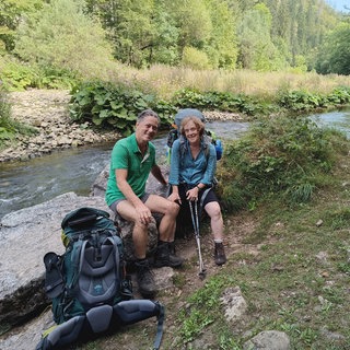Detlev Lindner und Marietheres Reul tragen Wanderkleidung. Sie sitzen auf einem Stein an einem Fluss. Neben ihnen steht ein großer Wanderrucksack. 
