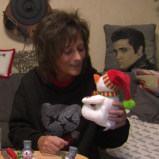 Ariane Weckbach sitzt auf einem Sofa und lächelt einen kleinen Schneemann aus Plüsch an, den sie in der Hand hält. Sie ist umgeben von Weihnachtsdekoration. 