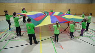 Eine Sportgruppe steht im Kreis und schwingt gemeinsam ein buntes Tuch. Alle tragen grüne Shirts.