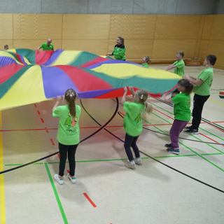 Eine Sportgruppe steht im Kreis und schwingt gemeinsam ein buntes Tuch. Alle tragen grüne Shirts.