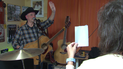 Jürgen Weckbach steht vor einem Mikrofon. Er hat eine Gitarre umgehängt und trägt einen Hut. Er hebt seine linke Hand und grüßt. Seine Frau Ariane steht vor ihm und hält einen Zettel in der Hand. 