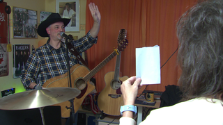 Jürgen Weckbach steht vor einem Mikrofon. Er hat eine Gitarre umgehängt und trägt einen Hut. Er hebt seine linke Hand und grüßt. Seine Frau Ariane steht vor ihm und hält einen Zettel in der Hand. 