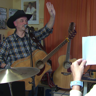 Jürgen Weckbach steht vor einem Mikrofon. Er hat eine Gitarre umgehängt und trägt einen Hut. Er hebt seine linke Hand und grüßt. Seine Frau Ariane steht vor ihm und hält einen Zettel in der Hand. 