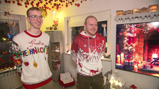 Zwei junge Männer stehen in Weihnachtspullovern in einem weihnachtlich geschmückten Raum. Sie lächeln. 
