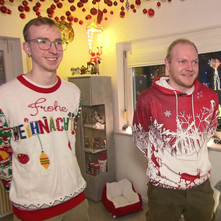 Zwei junge Männer stehen in Weihnachtspullovern in einem weihnachtlich geschmückten Raum. Sie lächeln. 