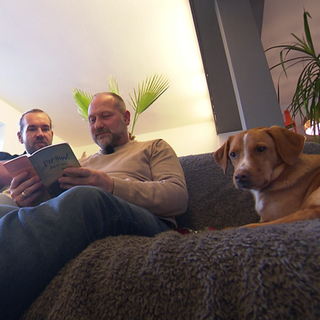 Steffen, sein Mann Christop und der Labrador-Golden Retriever Mischling Rudy sitzen zusammen auf einem Sofa. 