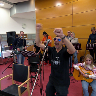 Eine Musikgruppe. Im Hintergrund stehen Männer und Frauen mit Instrumenten. Im Vordergrund ist ein Mann mit Sonnenbrille, der vor einem Mikrofon steht. Er streckt beide Arme in die Luft und hat den Mund weit geöffnet. 