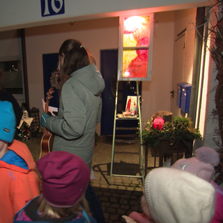Vor einem Haus stehen viele Kinder und einige Erwachsene. Ein erleuchtetes Fenster hängt über einer Leiter. Das Fenster ist bunt bemalt.