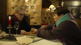 Sonja Faber-Schrecklein sitzt mit einem Mann an einem Tisch. Sie greifen beide zu einem Teller. Im Hintergrund ist ein heller Kachelofen.