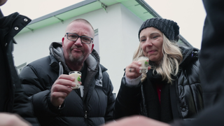 Zwei winterlich angezogene Menschen. Ein Mann und eine Frau, beide mit einem kleinen Glas in der Hand. 